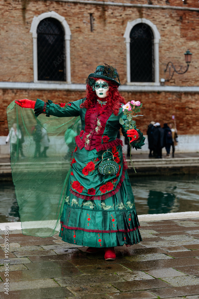 Naklejka premium Venice, Italy - February 09, 2026: Woman in elaborate green costume with mask and flowers poses during Venice Carnival near canal and historic brick buildings