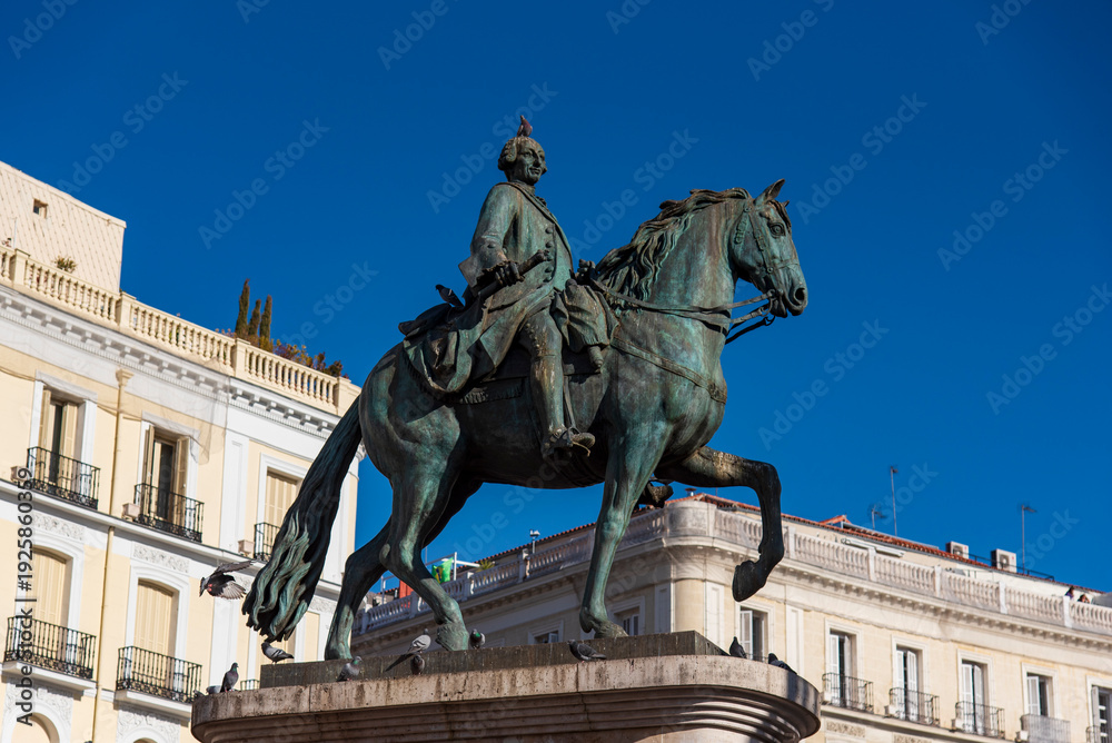Obraz premium Equestrian statue of King Philip III at Plaza Mayor in Madrid, Spain, historic landmark surrounded by colorful facades and towers under a clear blue sky.