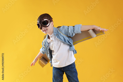 Bilde på lerret Happy little boy in goggles with cardboard plane wings playing pilot on yellow b