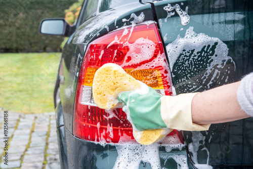 Woman's gloved hand scrubbing the tail light of a dark green car with a yellow sponge and white soap suds, performing car wash and exterior detailing for vehicle maintenance