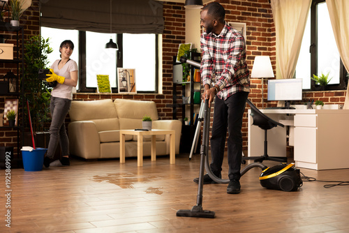 фотографія Smiling african american man vacuums hardwood floor while Caucasian wife wipes shelves in modern living room