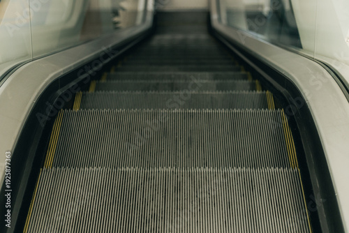 Close-up view of escalator steps leading downward, showcasing metal grooves and yellow safety lines, with glass panels on either side in a modern building interior