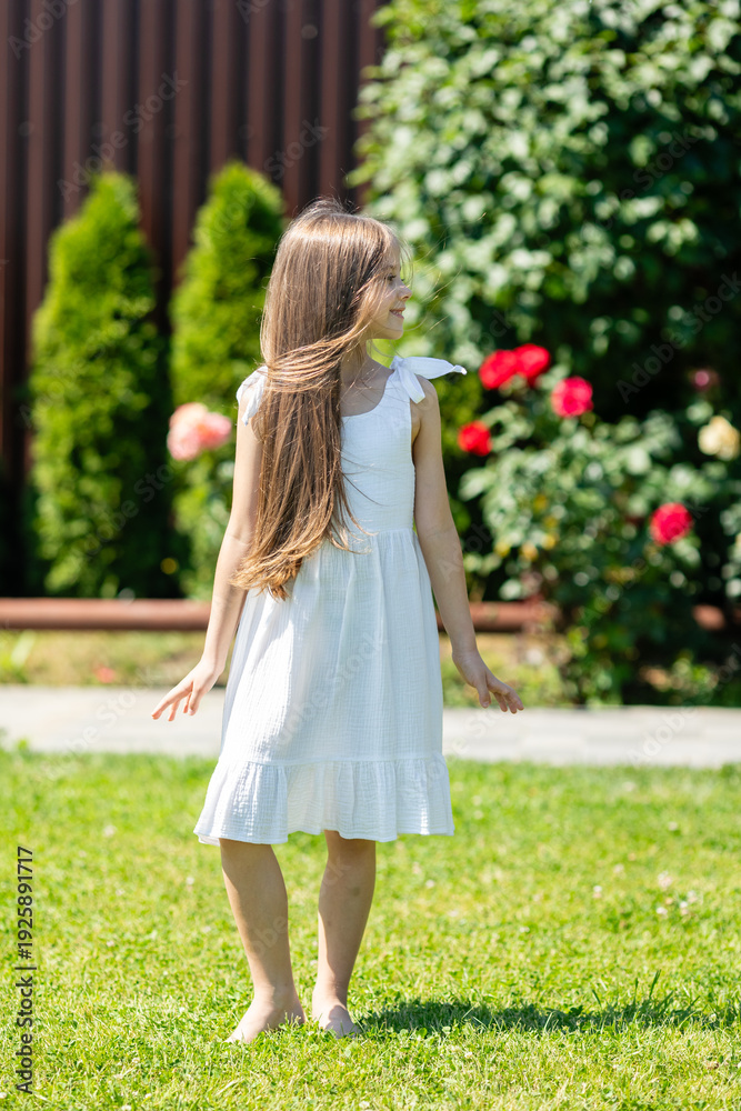 Obraz premium Cute little girl with long hair wearing a white dress stands barefoot on green grass in a garden with colorful flowers and shrubs in the background