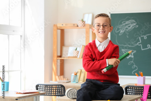 Cute little schoolboy with large pencil sitting on desk in classroom