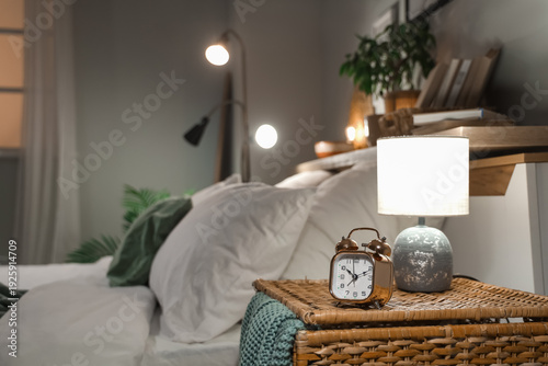 Tableau sur toile Cozy modern bedroom interior at night with bedside lamp, alarm clock, wicker nig