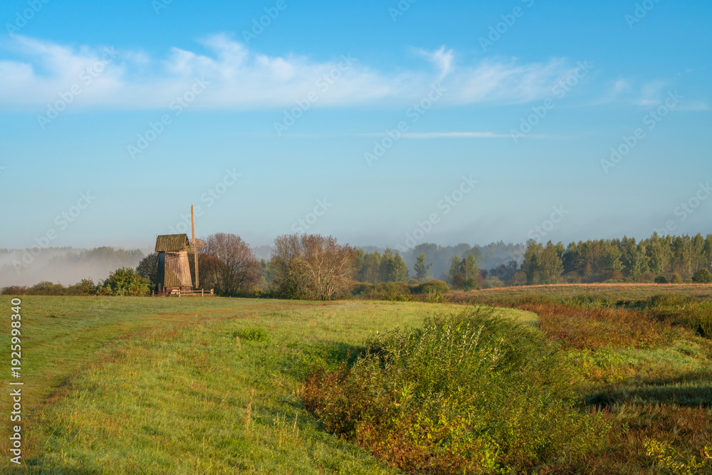 Fototapeta premium An ancient wooden windmill on the banks of the Sorot River on a foggy summer morning, Mikhailovskoye, Pushkinskiye Gory, Pskov region, Russia