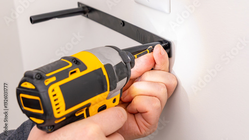 Canvas Print Mans hands operating yellow cordless drill attaching metal bracket to white wall with screw