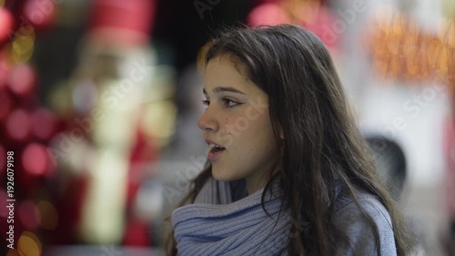 Wallpaper Mural A charming young girl with long dark hair and a light blue scarf smiles and observes her surroundings. Her joyful expressions reflect curiosity and happiness in a festive, blurred background. Torontodigital.ca