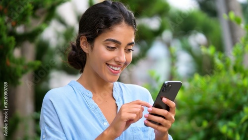 Woman is smiling while looking at her cell phone. She is holding the phone in her right hand and she is texting or browsing the internet. Concept of leisure and relaxation