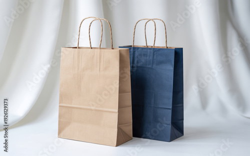 Two retail paper shopping bags, one brown kraft and one navy blue, stand side-by-side against a soft white fabric curtain backdrop, representing e-commerce, gift giving, sustainable packaging, and bou
