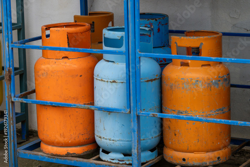 a blue metal storage rack containing several pressurized gas cylinders, specifically highlighting two bright orange propane tanks and one light blue cylinder with visible wear and rust on their surfac