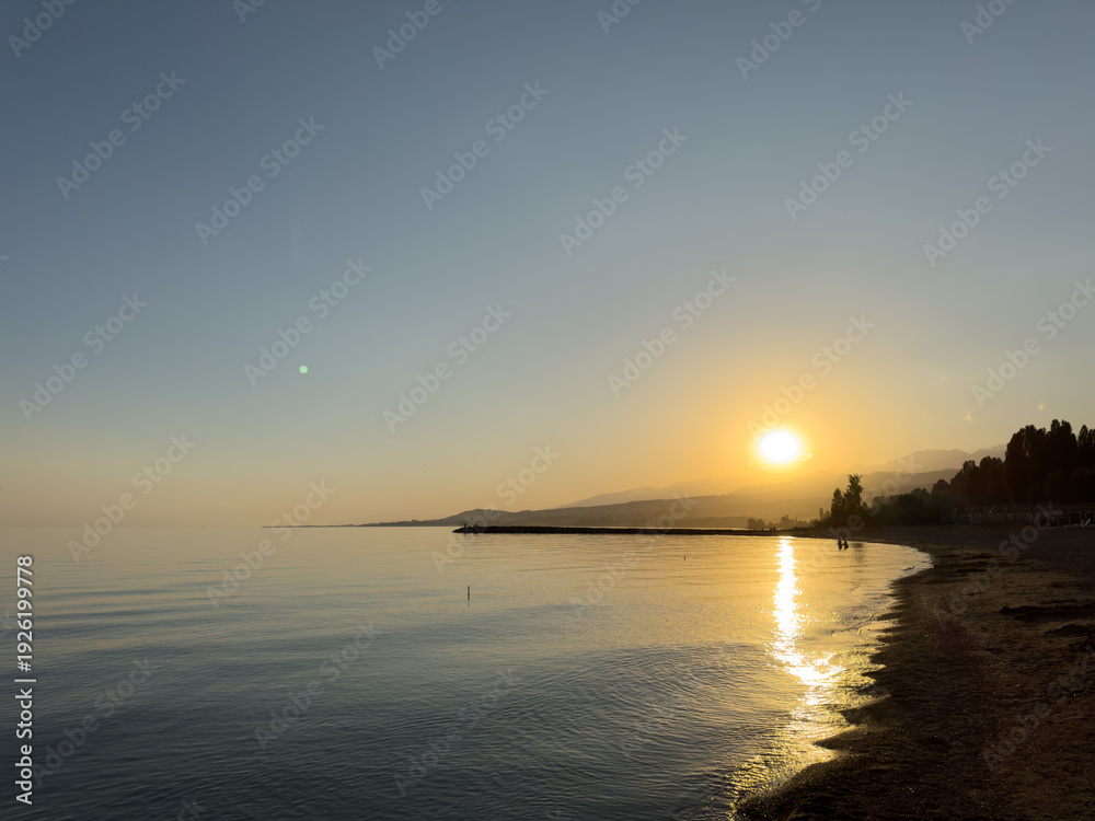 Obraz premium Scenic sunset over Issyk Kul Lake in Kyrgyzstan. The sun sets behind the mountains, casting a golden reflection across the water.