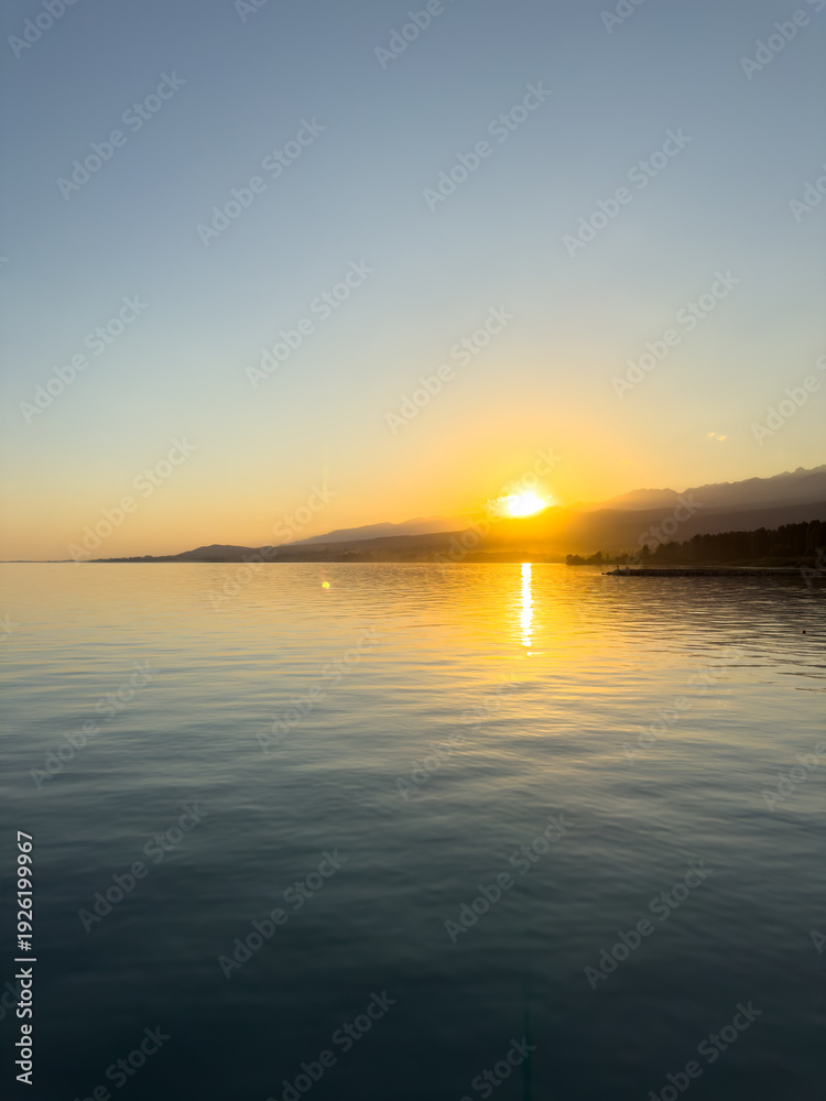 Obraz premium Scenic sunset over Issyk Kul Lake in Kyrgyzstan. The sun sets behind the mountains, casting a golden reflection across the water.