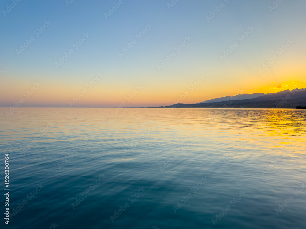 Obraz premium Scenic sunset over Issyk Kul Lake in Kyrgyzstan. The sun sets behind the mountains, casting a golden reflection across the water.