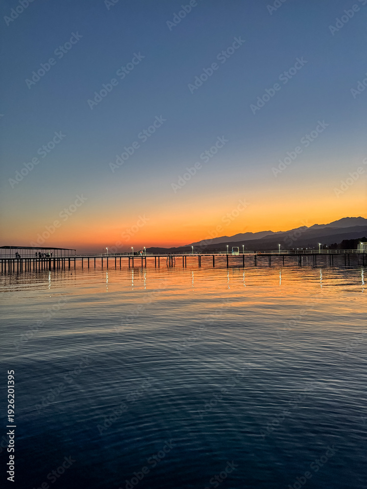 Naklejka premium Evening sunset at Issyk Kul Lake in Kyrgyzstan. Long illuminated pier stretching into calm water with mountains on horizon.
