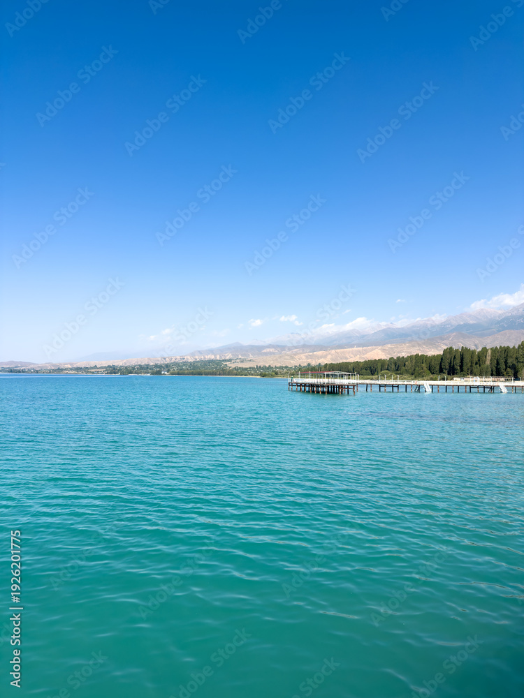 Fototapeta premium Issyk Kul Lake in Kyrgyzstan on a sunny day. Turquoise water, long pier and Tien Shan mountains on the horizon.
