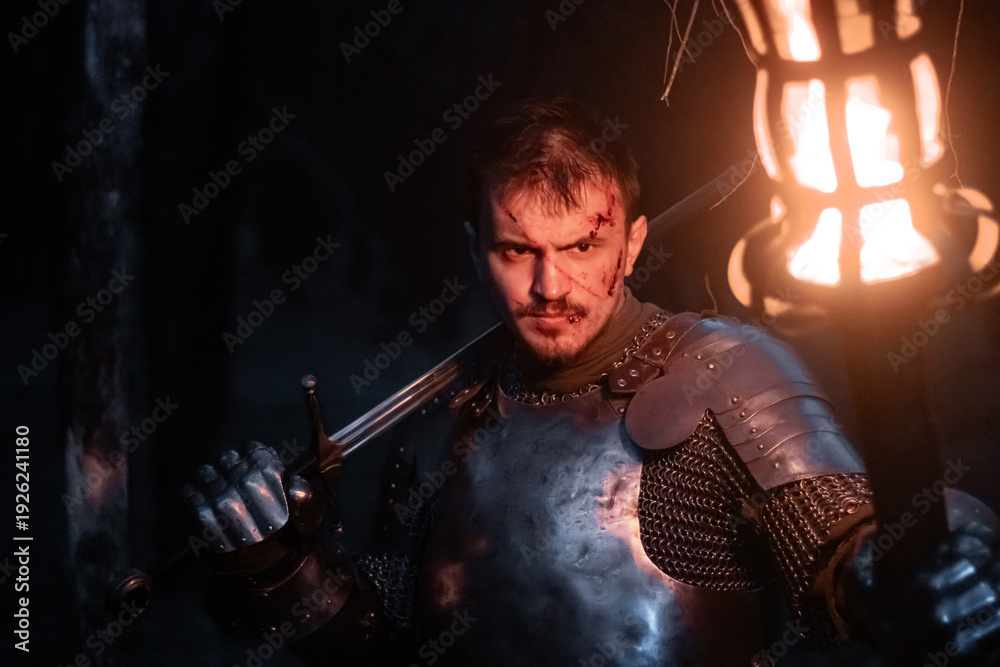 Obraz premium A knight in full armor, sword and torch in hand, walks through a gloomy night forest. A medieval warrior, bloodied and scarred, hunts and searches in the darkness.