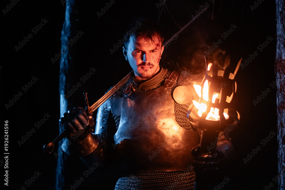 Obraz premium A knight in full armor, sword and torch in hand, walks through a gloomy night forest. A medieval warrior, bloodied and scarred, hunts and searches in the darkness.