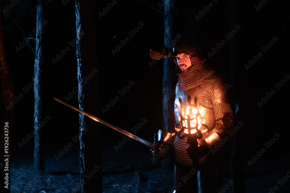 Obraz premium A knight in full armor, sword and torch in hand, walks through a gloomy night forest. A medieval warrior, bloodied and scarred, hunts and searches in the darkness.