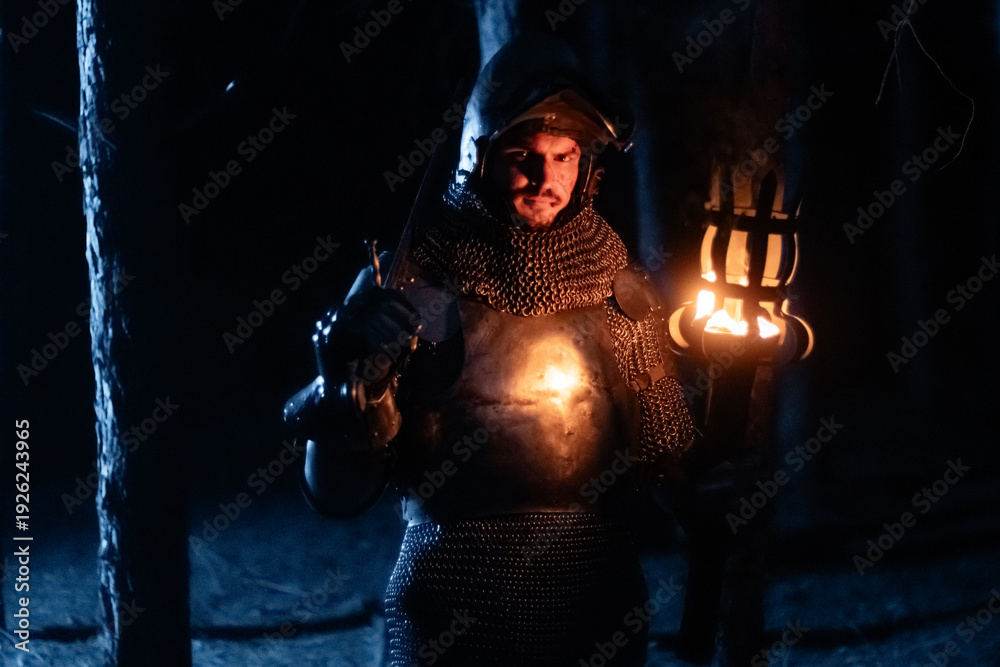 Obraz premium A knight in full armor, sword and torch in hand, walks through a gloomy night forest. A medieval warrior, bloodied and scarred, hunts and searches in the darkness.