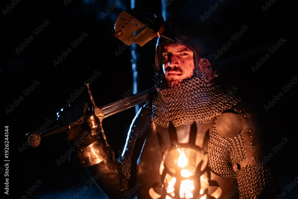 Obraz premium A knight in full armor, sword and torch in hand, walks through a gloomy night forest. A medieval warrior, bloodied and scarred, hunts and searches in the darkness.