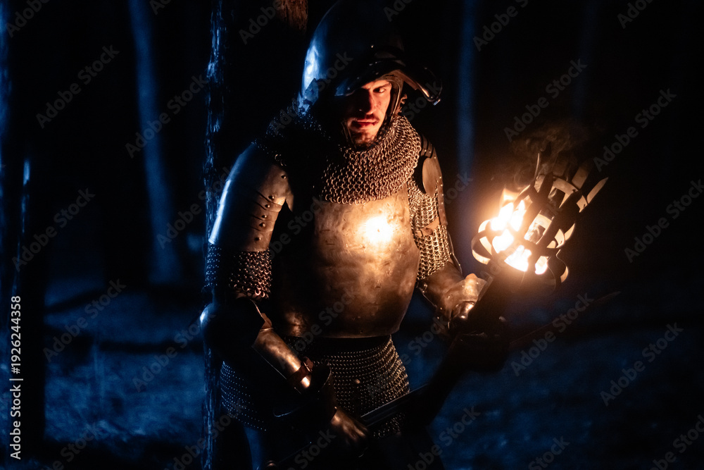 Obraz premium A knight in full armor, sword and torch in hand, walks through a gloomy night forest. A medieval warrior, bloodied and scarred, hunts and searches in the darkness.