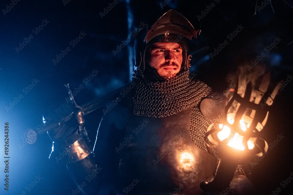 Obraz premium A knight in full armor, sword and torch in hand, walks through a gloomy night forest. A medieval warrior, bloodied and scarred, hunts and searches in the darkness.