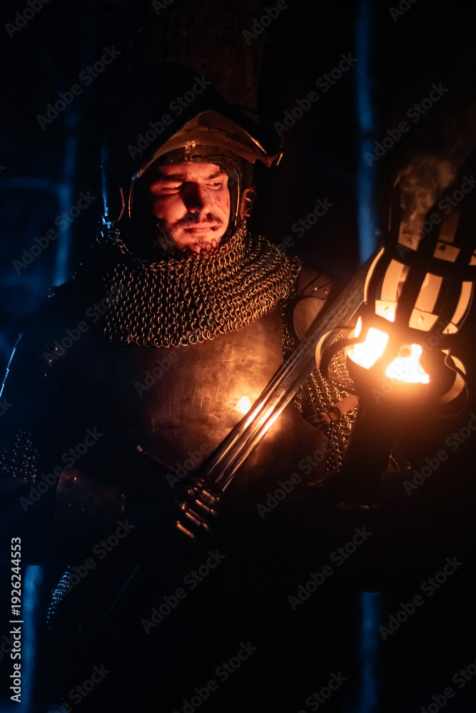 Obraz premium A knight in full armor, sword and torch in hand, walks through a gloomy night forest. A medieval warrior, bloodied and scarred, hunts and searches in the darkness.