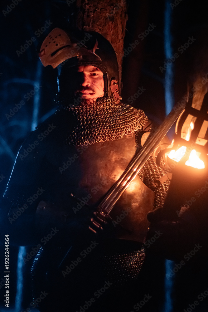 Obraz premium A knight in full armor, sword and torch in hand, walks through a gloomy night forest. A medieval warrior, bloodied and scarred, hunts and searches in the darkness.