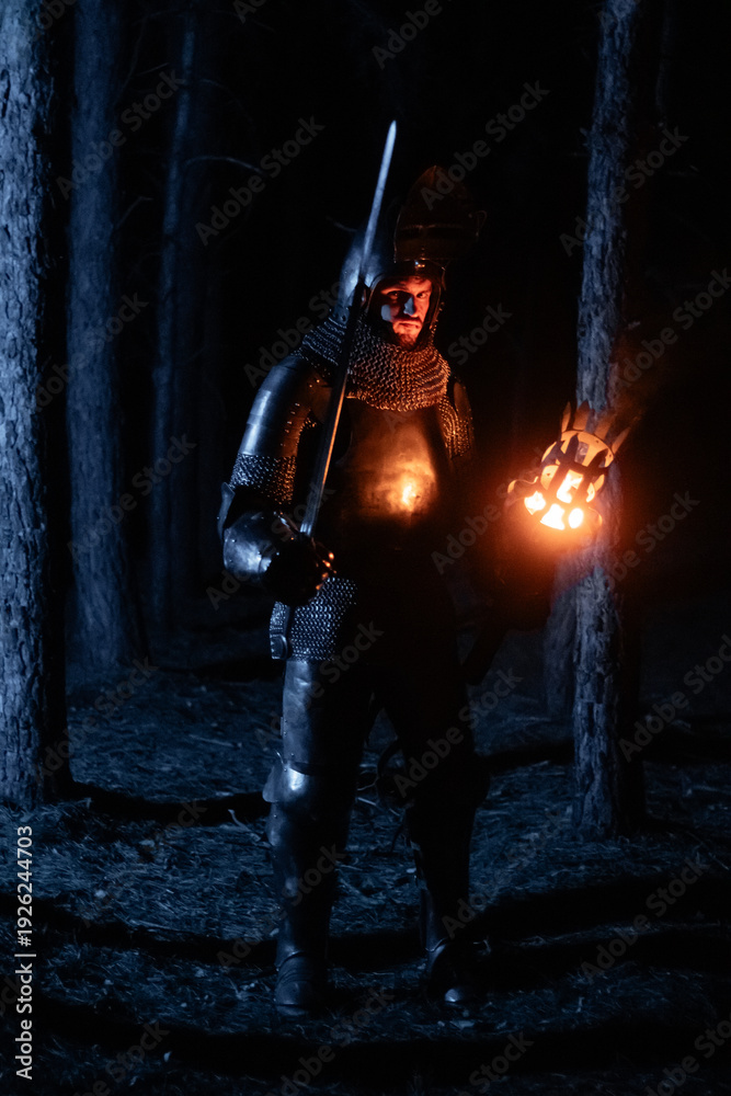 Obraz premium A knight in full armor, sword and torch in hand, walks through a gloomy night forest. A medieval warrior, bloodied and scarred, hunts and searches in the darkness.