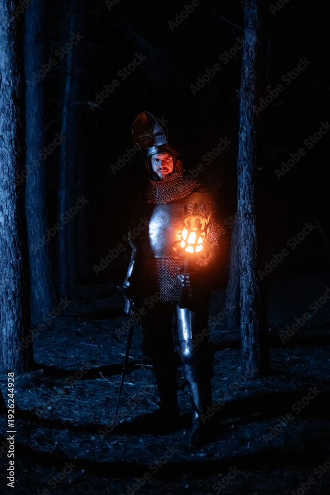 Obraz premium A knight in full armor, sword and torch in hand, walks through a gloomy night forest. A medieval warrior, bloodied and scarred, hunts and searches in the darkness.