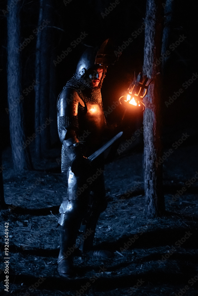 Obraz premium A knight in full armor, sword and torch in hand, walks through a gloomy night forest. A medieval warrior, bloodied and scarred, hunts and searches in the darkness.