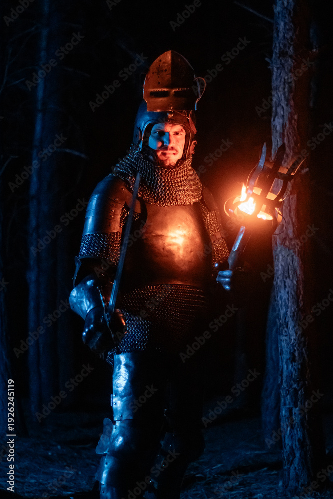 Obraz premium A knight in full armor, sword and torch in hand, walks through a gloomy night forest. A medieval warrior, bloodied and scarred, hunts and searches in the darkness.