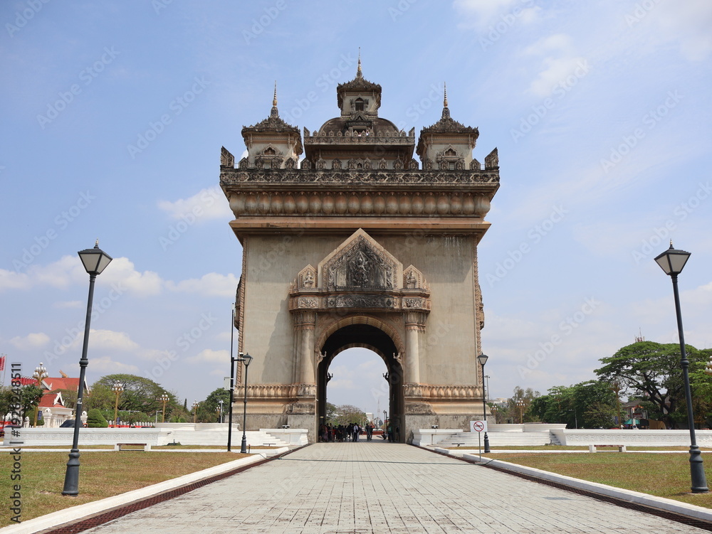 Fototapeta premium VIENTIANE, LAOS - FEBRUARY 25, 2026: The iconic Patuxay Victory Gate monument under a blue sky with tourists visiting.