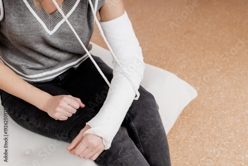 Close up of woman broken arm in white plaster cast gypsum orthopedic sling immobilization sitting on sofa at home during fracture injury recovery rehabilitation treatment showing patient daily life