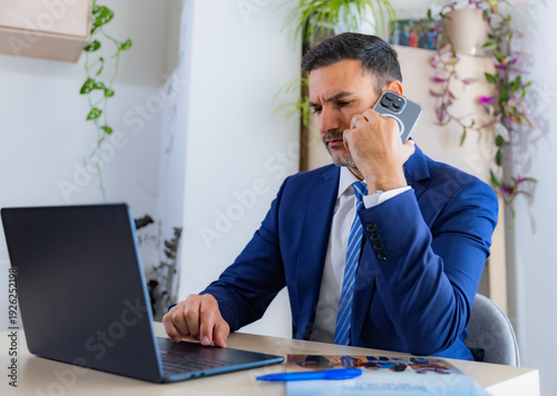 Billede på lærred Young entrepreneur talking anxiously on his cell phone while working on his laptop