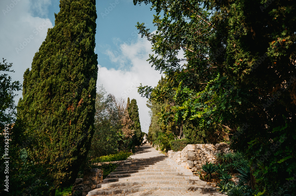 Fototapeta premium Sunlit El Calvari steps surrounded by greenery in Pollença, Mallorca