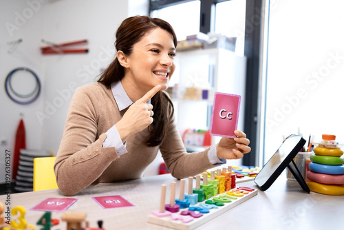 Female speech therapist on a video call holding an alphabet flashcard c while demonstrating mouth articulation for a child's remote phonics and language development lesson