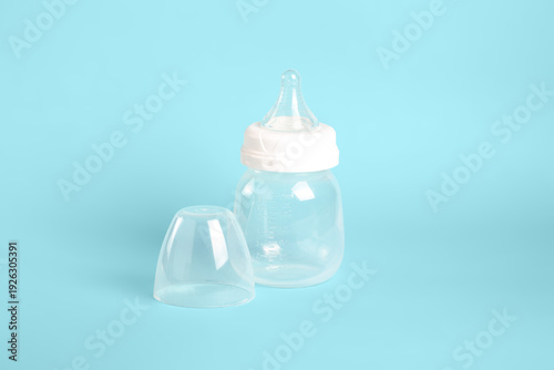 A clear milk bottle sits on a light blue background. The bottle has a white lid placed beside it. The scene shows a simple setup for feeding infants or young children.