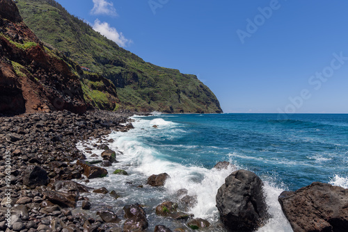 Rocky Atlantic coastline near Porto Moniz on Madeira Island Portugal, dark volcanic stones meet rolling surf and blue water beneath wide clear summer sky