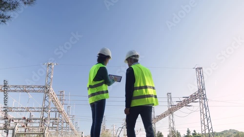 Electrical engineers with a digital tablet discussing maintenance and inspecting an electric power grid substation, ensuring safety and efficiency in energy distribution
