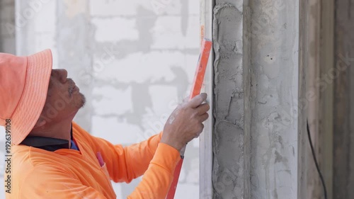 Construction worker checking wall alignment with spirit level during plastering process. Interior renovation, precision measurement and professional building concept.