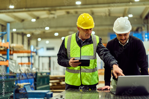Manufacturing Supervisors in Hard Hats Reviewing Production Plans on a Factory Floor Laptop and Tablet