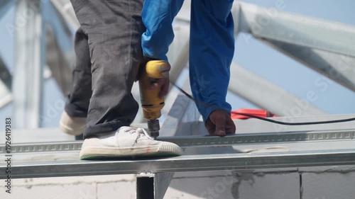 Construction worker using electric drill to fasten steel roof frame at building site. Industrial installation, structural engineering and roofing project concept.