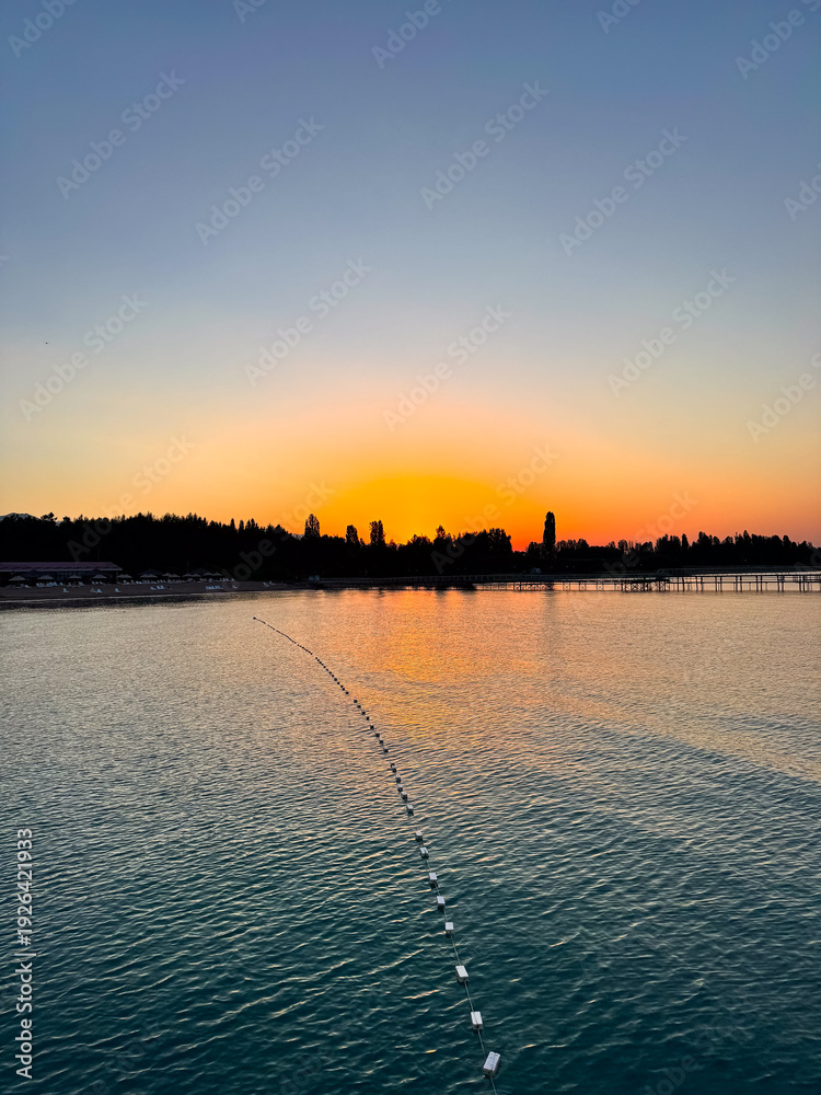 Fototapeta premium Early sunrise at Issyk Kul Lake in Kyrgyzstan. Warm golden sky reflecting in calm water with silhouettes of trees and pier on horizon.
