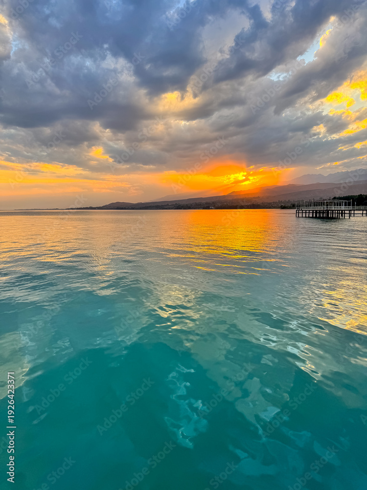 Fototapeta premium Sunset at Issyk Kul Lake in Kyrgyzstan. Bright sun breaking through clouds reflecting in clear turquoise water.
