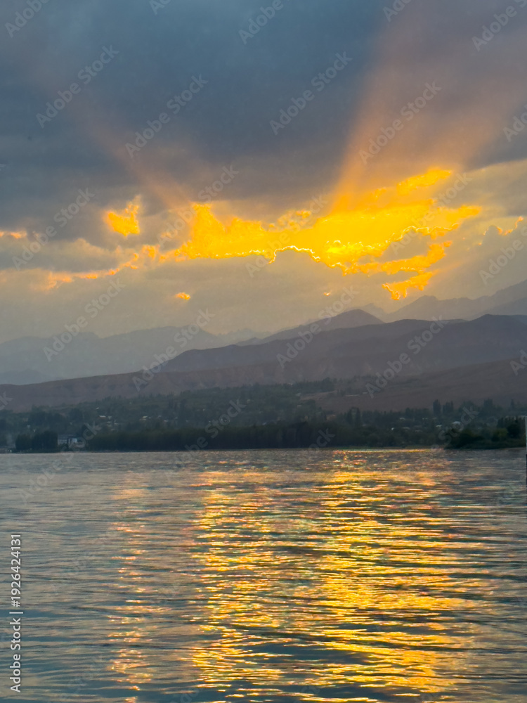Fototapeta premium Sunset at Issyk Kul Lake in Kyrgyzstan. Bright sun breaking through clouds reflecting in clear turquoise water.