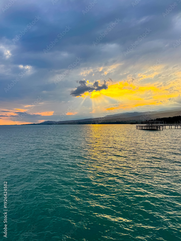Fototapeta premium Sunset at Issyk Kul Lake in Kyrgyzstan. Bright sun breaking through clouds reflecting in clear turquoise water.