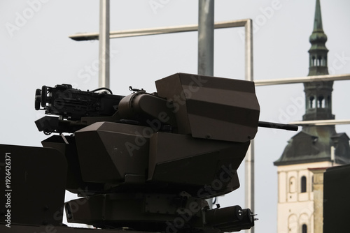 Modern remote controlled weapon station with heavy machine gun mounted on armored vehicle turret against urban city background and cathedral