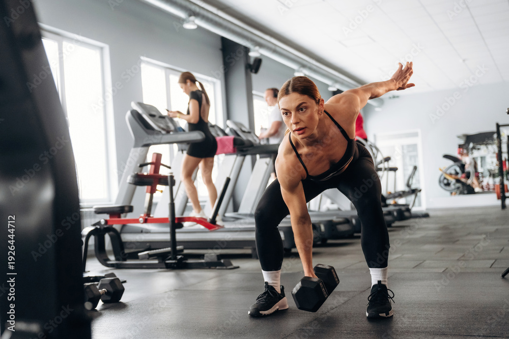 Fototapeta premium Female fitness enthusiast performing a bent-over dumbbell row exercise in a modern gym with treadmills and exercise equipment in the background